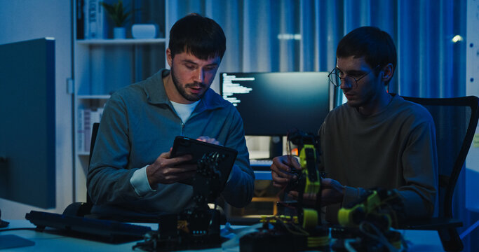 Team adult male engineer collaborate on robotic hand project using AI code solutions machine learning at tech workspace night. Artificial intelligence interface, collaboration prototype development.
