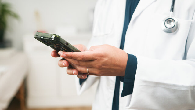 Doctor Using Smartphone In Clinic With Stethoscope Draped Over White Coat For Medical Communication