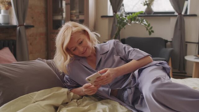 Zoom in shot of mature Caucasian woman with blond hair wearing comfortable pyjamas relaxing on bed in morning surfing social net in Internet on smartphone
