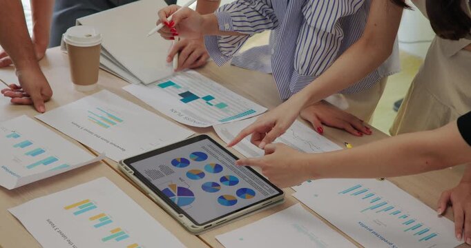 Close-up view of diverse young business team collaborating with digital tablet and printed charts. Hands pointing at colorful graphs and analytics during a strategy planning session at office.