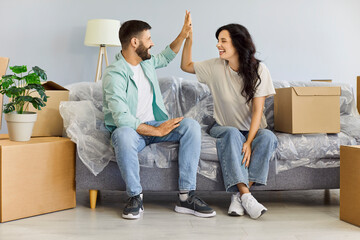Happy couple celebrating first moving day with with high five and laughter, cheerful people sitting on sofa among cardboard boxes. Young man and woman laughing with love, planning unpacking together