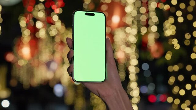 Closeup view of woman hand holds modern smartphone with green chromakey screen against bokeh lights outdoors. Female shows mockup screen of mobile phone in festive evening town