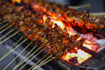 Chicken Satay or Sate Ayam skewers coated in thick peanut sauce, being grilled over glowing charcoal embers on a street vendor's portable grill at night.