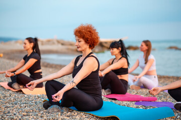 Meditation class at sea coast. Group of Caucasian women sitting on sports mats on pebble beach. Yoga outdoor training