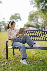 Obraz premium Woman sitting on a park bench wearing headphones and smiling, using a digital tablet for entertainment, education, or communication while enjoying free time outdoors