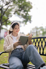 Young Asian woman sitting on a park bench relaxing and enjoying leisure time, reading a book and listening to music using wireless headphones outdoors