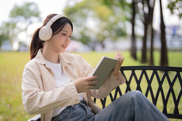 Obraz premium Smiling Asian woman sitting on a park bench, enjoying music with headphones and interacting with a digital tablet, embracing leisure and technology in nature