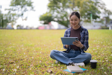 Young Asian woman sitting cross-legged on green grass in a park, smiling while engaging in online learning and remote education on a digital tablet with a takeaway coffee and books beside her