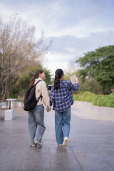 Obraz premium Two young women, dressed in casual clothes, walking together and holding hands on a paved path in a park-like outdoor setting, sharing a moment of friendship