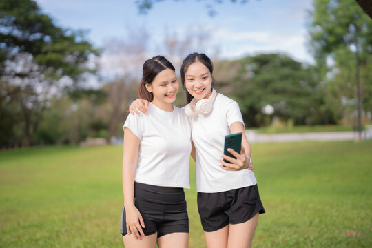 Two young Asian women, best friends, happily taking a selfie together with a smartphone in a green park, embracing friendship and enjoying active life