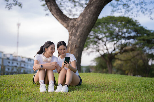 Two young Asian women sitting on grass in an urban park, smiling and sharing content on a smartphone while enjoying leisure time, nature, and friendly connection