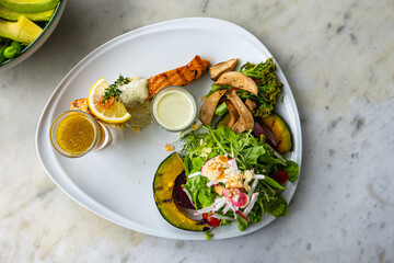 Top view of roasted salmon steak cooked with various vegetable salad served on white ceramic plate with lemon cream sauce on marble top table.