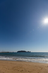 Sun-drenched sandy beach meets sparkling turquoise ocean with distant island and bright sunburst sky