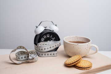 Сup of tea with cookies and an alarm clock wrapped in a measuring tape against a neutral background. Conceptual ideas for time management, healthy living, diet, and balancing a morning routine.