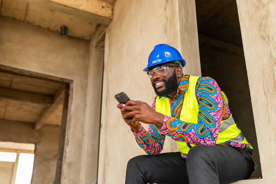 Smiling African construction engineer in hard hat and safety vest using smartphone for video call, shopping, digital banking, social media, internet browsing