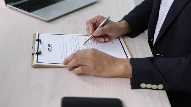 Businessman wearing a suit sitting at an office desk and filling a resume form placed on a clipboard with a pen, laptop and smartphone on the desk.