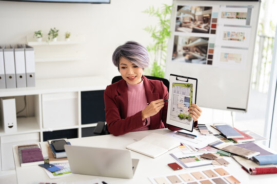 Interior designer talking with client on video call, showing floor plan blueprint of residence and pantone. Asian architect in studio office, looking at laptop computer display with online conference.