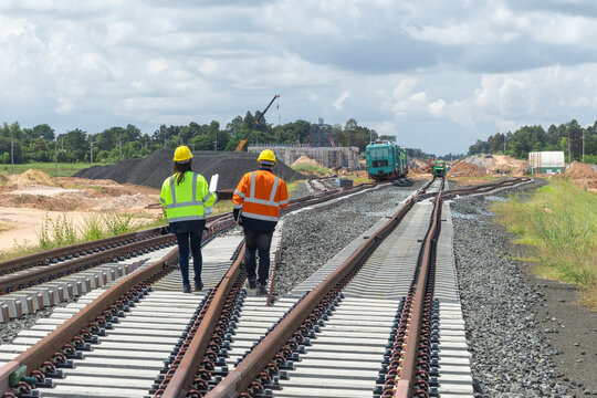 Two Asian engineers or workers wearing safety gear and hard hats, inspect railroad track construction at station. Concept for elevated railway and double-track railway construction in Thailand.