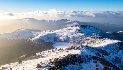 A sunlit aerial view reveals a snow-covered mountain range under a clear sky. Layers of snow cover peaks, revealing a frozen lake below