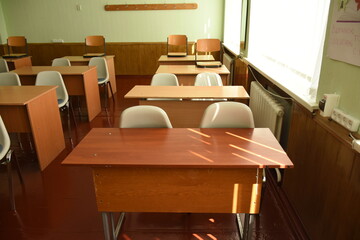 Classroom interior with brown student desks for two and mixed chairs — white and brown. In the background, there is a coat hanging area along the wall. Bright learning environment with wooden furnitur