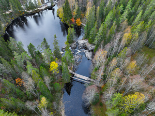 Mid-level aerial view of a dark river channel with whitewater rapids and a wooden bridge, bordered by tall pine and bare deciduous trees in autumn