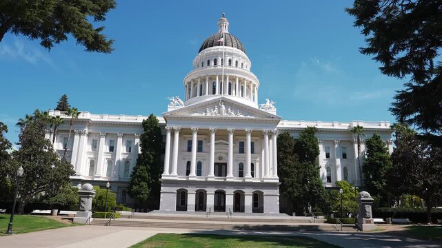 Close up shows the California State Capitol in Sacramento. The camera slowly pans or advances, revealing the dome, columns, pediment, steps, and Capitol Park trees in bright midday light.