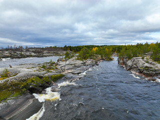 View of fast-moving, white-capped water flowing through a channel of gray, moss-covered granite rock formations with forest in the background