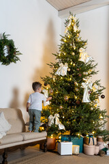 Young boy decorating a beautifully lit Christmas tree from a sofa in a cozy living room
