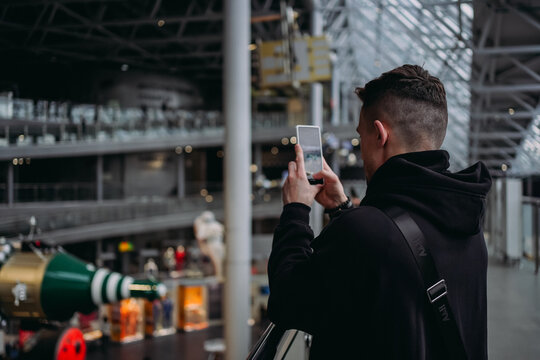Young man taking photo with smartphone inside modern museum, candid rear-view portrait with steel and glass architecture and soft natural light