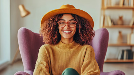 Cheerful young woman with vibrant smile and curly hair, wearing mustard yellow hat and glasses, comfortably seated in cozy purple armchair within bright indoor setting.