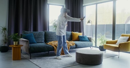 Elderly woman in virtual reality headset playing video game at home - Powered by Adobe