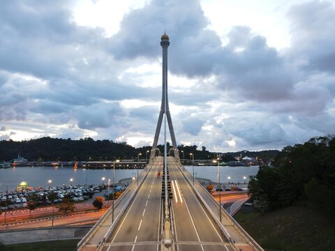 Aerial drone view of RIPAS Bridge in Bandar Seri Begawan, Brunei, at dusk. The cable-stayed bridge glows with lights over the river, framed by dramatic clouds and scenic city surroundings.