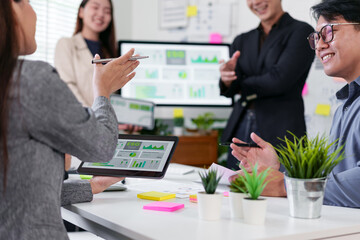 Business team meeting presenting sustainability charts on tablet and monitor, collaborative discussion with digital reports and smiling colleagues