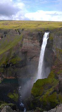 Haifoss plunges into a mossed canyon as mist and a faint rainbow form. Granni sits opposite. Aerial vertical view shows basalt columns and a turquoise pool below.