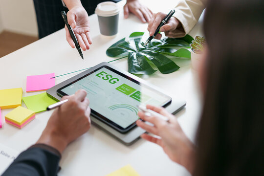 ESG report tablet surrounded by hands, pens, sticky notes and tropical leaf conveying collaborative sustainable business planning with focused