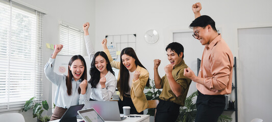 Young diverse coworkers celebrating team success around laptop with joyful expressions and raised fists in modern office