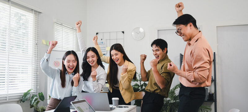 Young diverse coworkers celebrating team success around laptop with joyful expressions and raised fists in modern office - Powered by Adobe