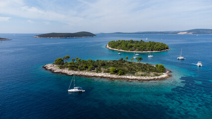 Obraz premium Aerial view of small green islands surrounded by deep blue sea with yachts and sailboats anchored near the rocky shore on a sunny day