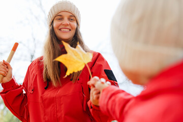 A cheerful woman wearing a red jacket and hat shares a vibrant yellow leaf with a young child during a sunny autumn day in the park