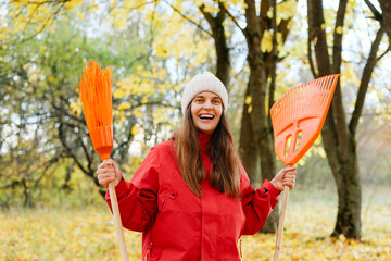 A cheerful woman is raking fallen leaves in a park filled with yellow trees, wearing a red jacket and a warm hat during the fall season