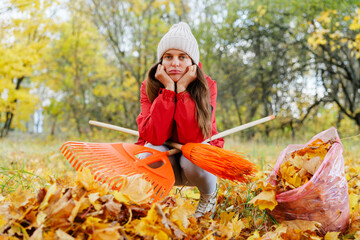 A woman in a red jacket and knitted hat sits among yellow leaves, tired while raking in a colorful autumn setting