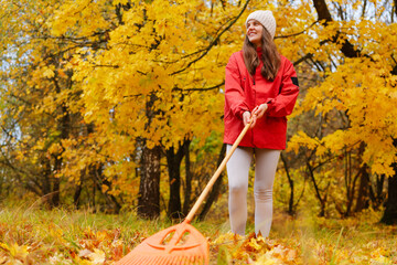 A woman in a red coat and hat is raking leaves in a vibrant, yellow autumn park filled with trees