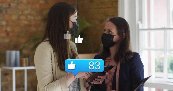 Two women wearing blazers, masks checking smartphone and tablet in brick office, with media icons