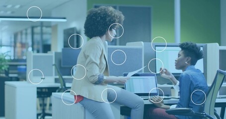 Two women in suits discussing printed reports while pointing at laptop charts in open-plan office