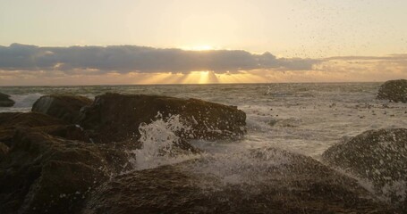 Crashing ocean waves spraying sea spray on jagged shore rocks, with sun rising behind low clouds