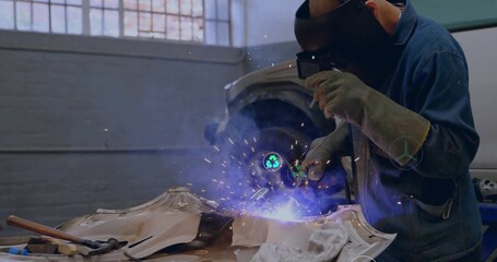 Welding technician directing torch at car body in workshop, wearing helmet, with sparks, copy space