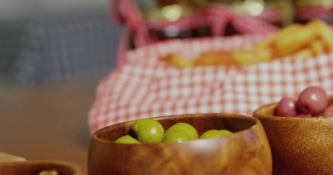 Displaying wooden bowl holding green olives on dining table, with purple olives and picnic basket