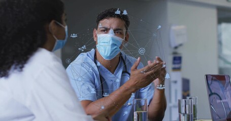 Gesturing doctor wearing scrubs discussing case in hospital office, with digital network overlay