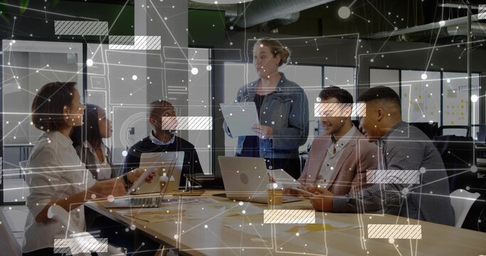 Collaborating team reviewing project data around table in meeting room, with laptops and tablets
