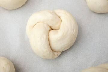 Top view of bread knot on baking sheet with paper. Raw bread dough knot rising or waiting to be baked. Traditional Swiss bread recipe called Zopf, Butterzopf or challah. Selective focus.